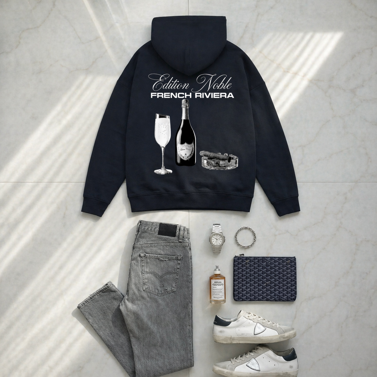 Black hoodie with 'Edition Noble French Riviera' design, paired with jeans, sneakers, a watch, and a clutch on a light gray floor.