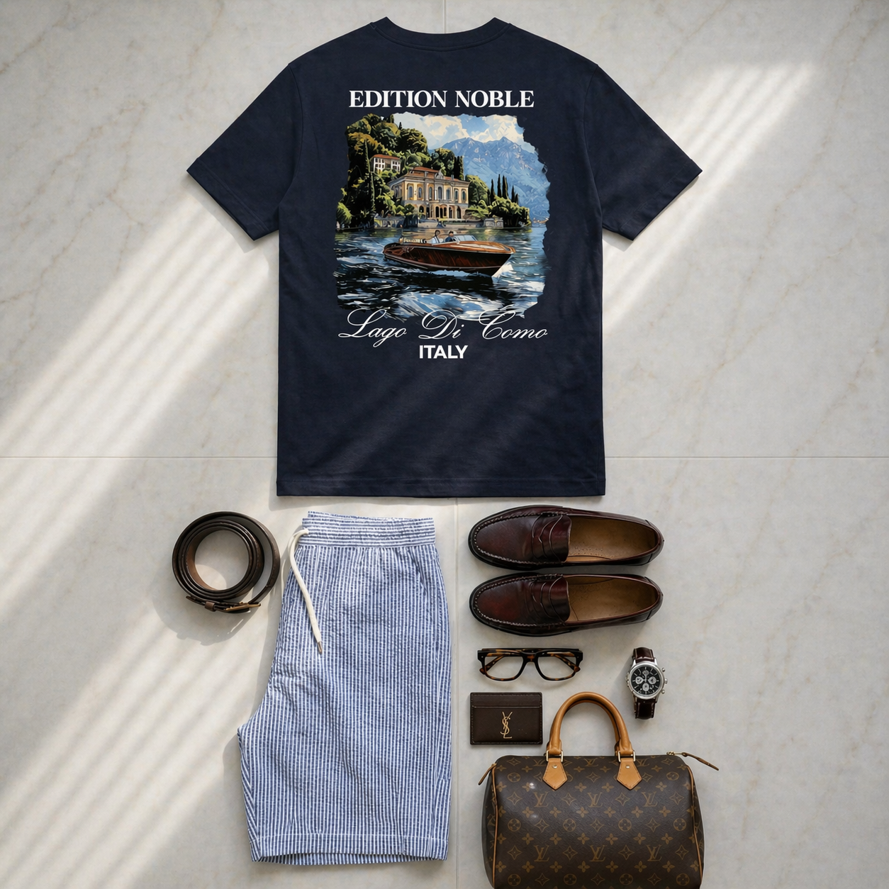Navy t-shirt with scenic print, shorts, shoes, glasses, wallet, handbag, and watch on a light gray surface.
