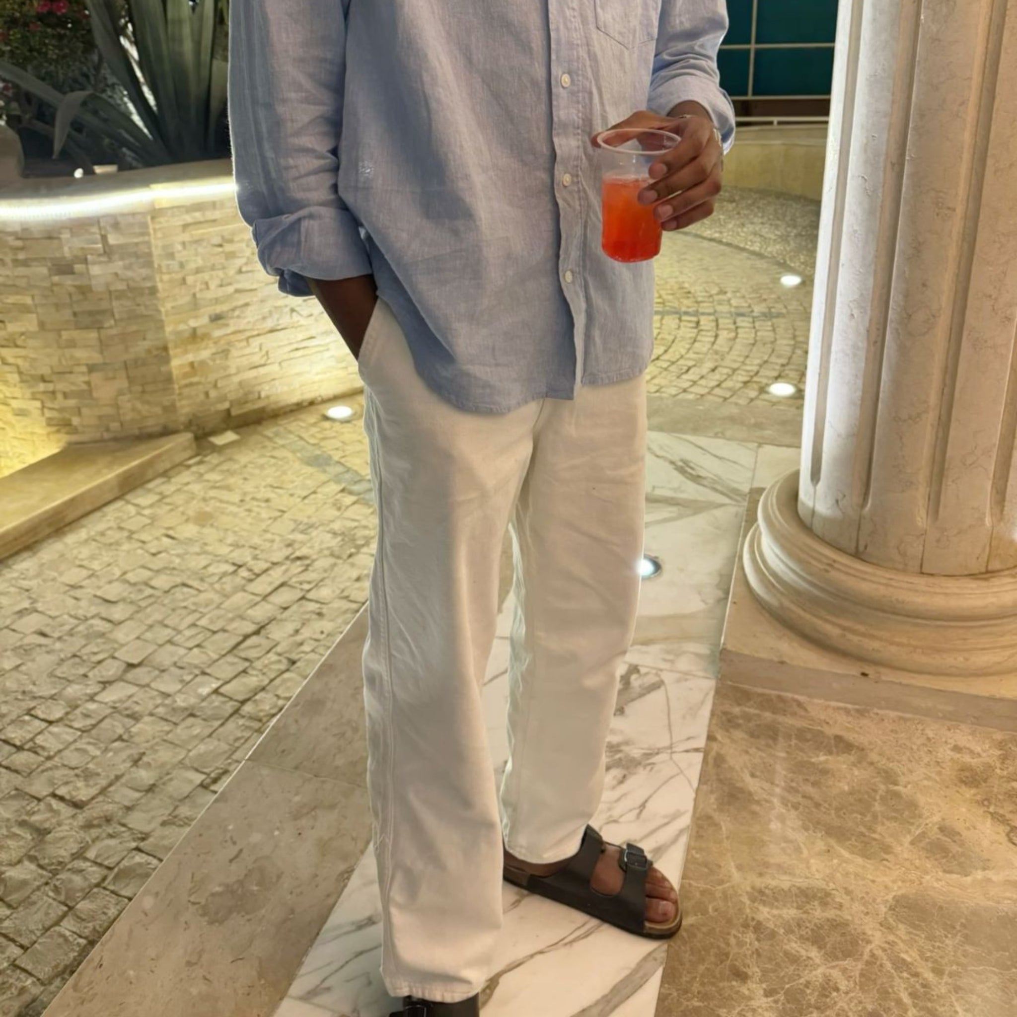 A person standing indoors near a marble column and tiled floor, wearing a light blue button-up shirt and white pants while holding a clear cup with an orange drink.
