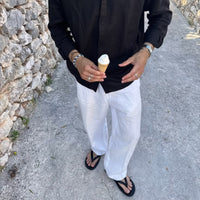 A person standing outdoors on a stone path wearing a black long-sleeve button-up shirt, white wide-leg pants, and black flip-flops, while holding an ice cream cone.